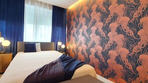 Une chambre à coucher avec un lit, deux tables de chevet avec des lampes, une fenêtre avec des rideaux et un mur orné d’un motif floral.
