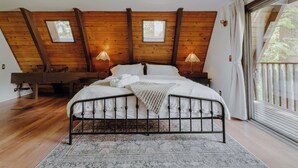 Quarto com teto de madeira, cama com roupa de cama branca, duas luminárias de cabeceira e uma grande janela com cortinas.