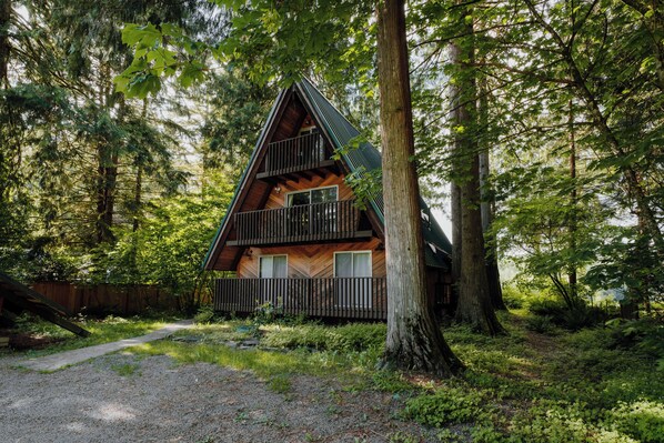 A cabin with a wooden exterior, a balcony, and a natural forest setting. - Spacious North Cascades Riverside A-Frame w/ Mt Baker Views - Dog-Friendly (Concrete)