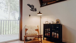 Quarto com piso de madeira, uma cadeira com manta de pele branca, uma luminária, uma estante e uma janela com persianas.