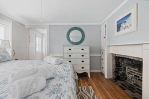 Une chambre à coucher comprenant un lit, une commode, un miroir, une cheminée et un tableau encadré accroché au mur.