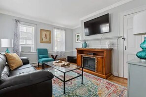 Un salon avec un canapé en cuir noir, un fauteuil turquoise, une table basse en verre, une cheminée et un téléviseur à écran plat fixé au-dessus de la cheminée.