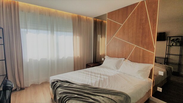 Une chambre d’hôtel moderne avec un grand lit, une tête de lit en bois ornée de motifs géométriques, une table de chevet et une grande fenêtre avec des rideaux légers.