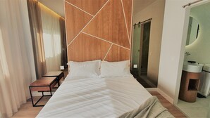 Une chambre d’hôtel moderne avec un grand lit, une t ête de lit en bois, une table de chevet, un banc et une salle de bain équipée d’un miroir et d’un lavabo.