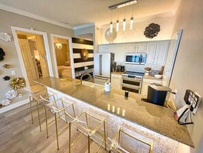 A modern kitchen with a granite countertop, stainless steel appliances, and a bar area with gold chairs. - Exquisite Beach Condo, Comfortable, Luxurious, Beachfront, Free Beach Service (Panama City Beach)