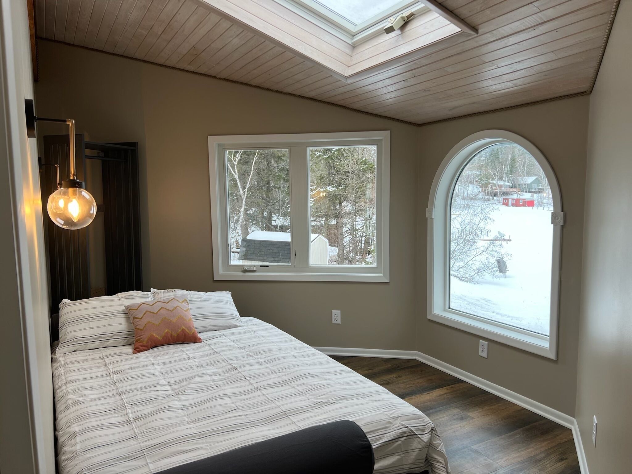 A bedroom with a bed, two windows, a skylight, and a hanging light fixture.