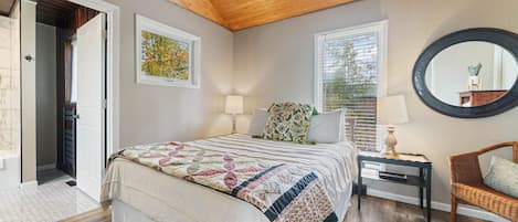 A bedroom with a wooden ceiling, a bed with a patterned quilt, a nightstand with a lamp, a wicker chair, and a round mirror on the wall.