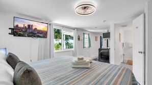 Una habitación de hotel moderna con una cama grande, un televisor de pantalla plana montado en la pared, un ventilador de techo y vistas de la ciudad a través de la ventana.