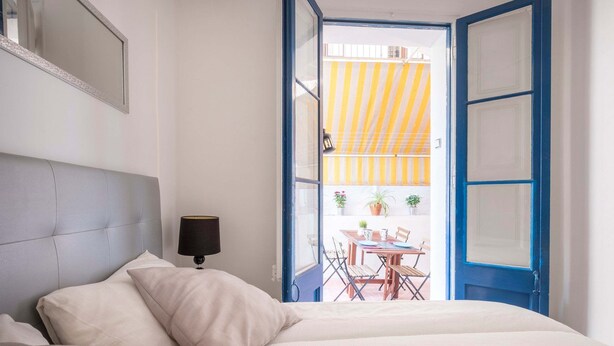 A bedroom with a bed, a bedside lamp, and a view of an outdoor dining area through an open door. - Apartment (Barcelona)