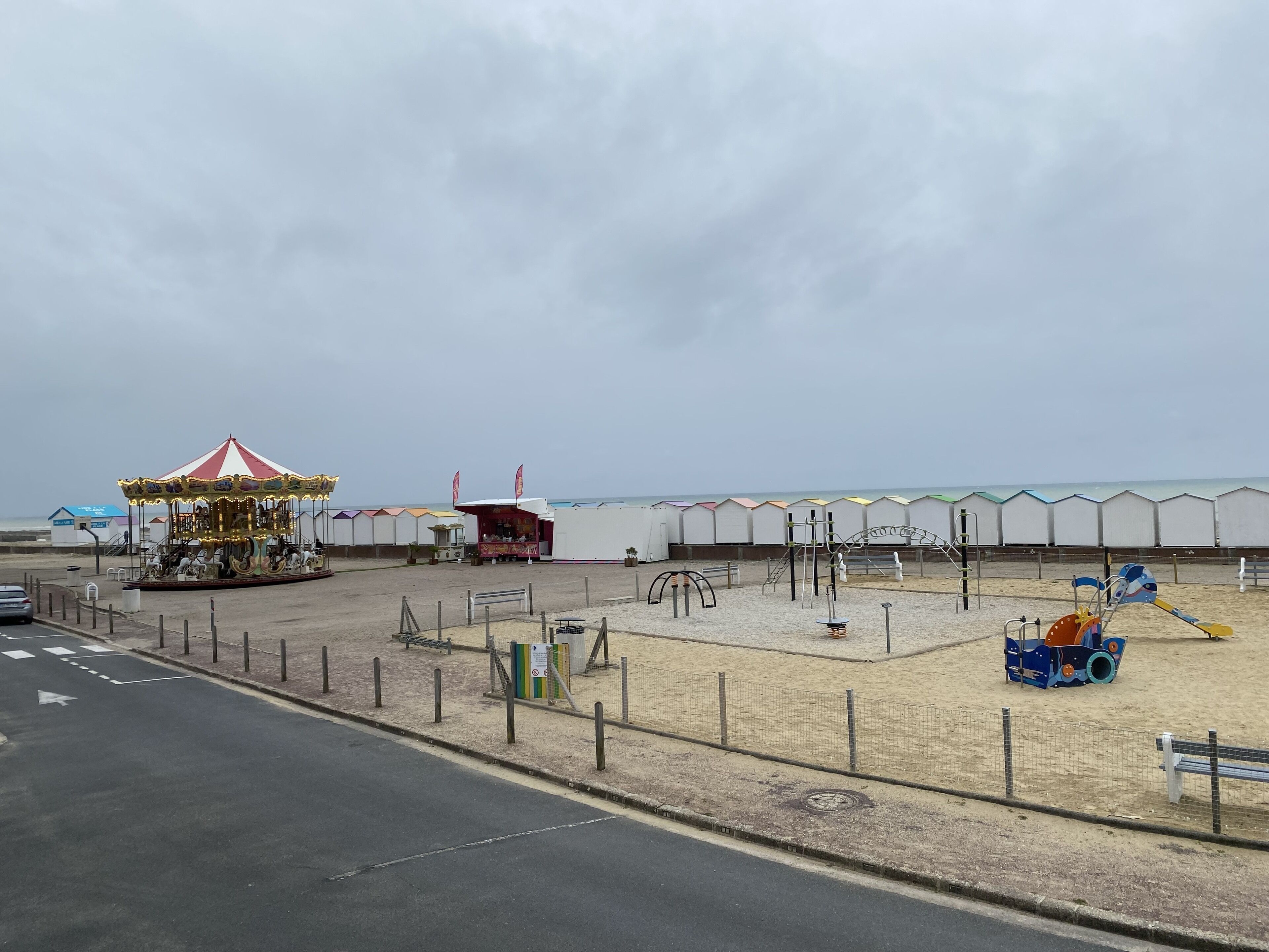 Un espace de loisirs situé au bord de la plage, comprenant un carrousel, des jeux pour enfants et une zone sablonne délimitée par une clôture.