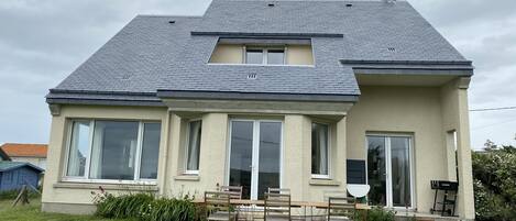 Ein zweistöckiges Haus mit geneigtem Dach, großen Fenstern und einer Veranda mit Tisch und Stühlen.