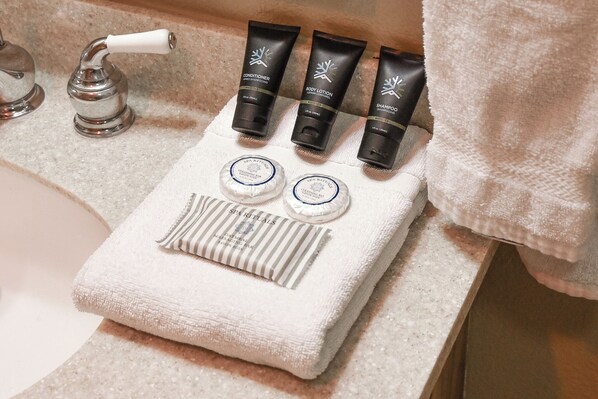 Kylpyhuoneessa on pyyhe, kaksi pulloa vartalovoideita ja yksi shampoo-tuubi, jotka ovat kaikki asetettuina pesualtaan reunalle.