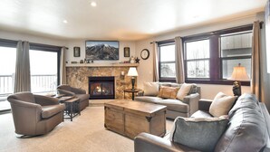 A spacious living room with a stone fireplace, leather sofas, a coffee table, and large windows offering a view of the outdoors. - Park Place Peak 10 Condo: Walk To Lift, Main Street (Breckenridge)