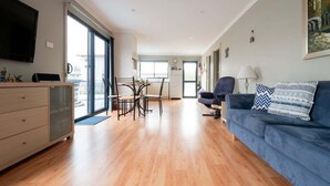 A modern living room with a blue sofa, a dining area with a table and chairs, and a kitchen in the background. - Cosy Coastal Retreat in Stieglitz with Water Views (Stieglitz)