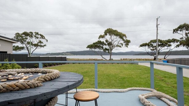 An outdoor deck with a round table and two stools, surrounded by rope barriers, overlooking a grassy area with trees and a distant view of hills. - Cosy Coastal Retreat in Stieglitz with Water Views (Stieglitz)