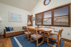 Uma área de refeições com mesa e cadeiras de madeira, tapete azul, sofá com almofadas e janela com persianas.