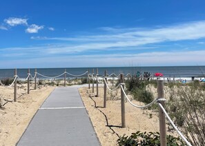Uma trilha de praia que leva ao oceano, com uma barreira de corda, areia e céu claro.