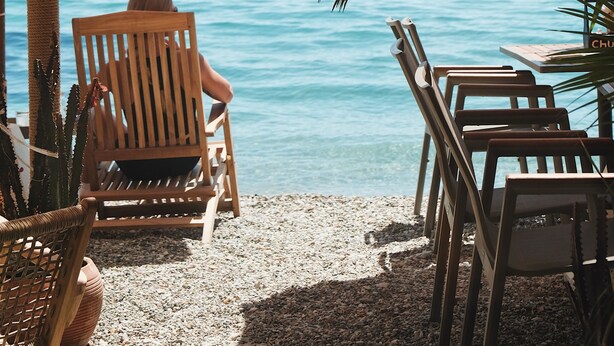 Ένας χώρος με καθίσματα δίπλα στην παραλία, με ξύλινα καθίσματα και τραπέζια, μια ομπρέλα με καλάμι και θέα στη θάλασσα, με ένα πλοίο να διακρίνεται στο βάθος.