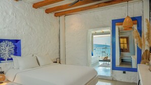 Ένα υπνοδωμάτιο με ένα μεγάλο κρεβάτι, έναν λευκό τοίχο με μια μπλε διακοσμητική λεπτομέρεια, ένα φωτιστικό από μπαμπού και θέα στη θάλασσα μέσα από μια ανοιχτή πόρτα.