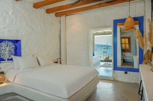 Una camera da letto con un letto grande, una parete bianca con un dettaglio blu, una lampada in vimini e vista sul mare attraverso una porta aperta.