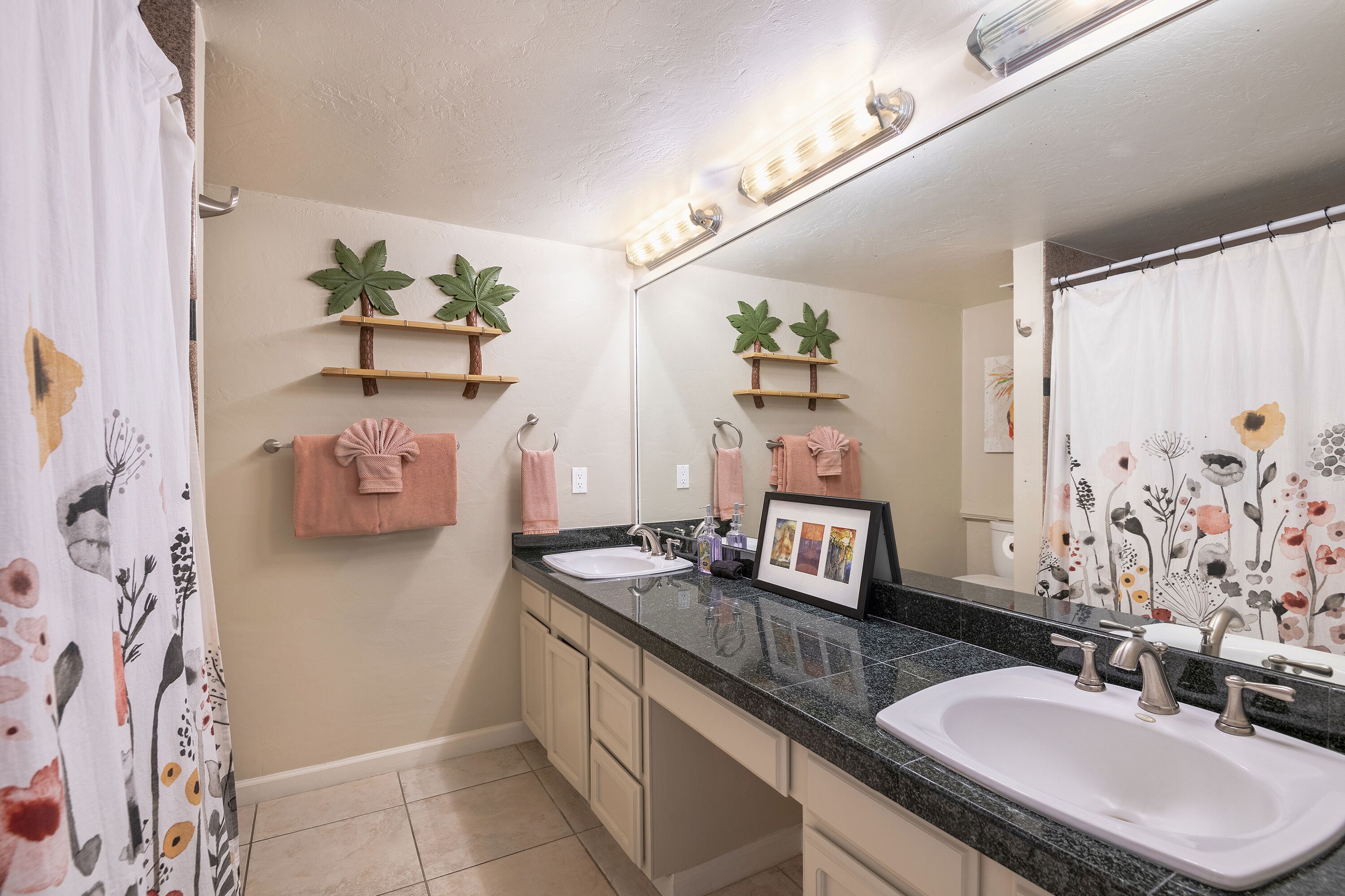 A bathroom with a double sink, vanity, and a mirror. There are decorative plants on the wall and a towel rack with towels.