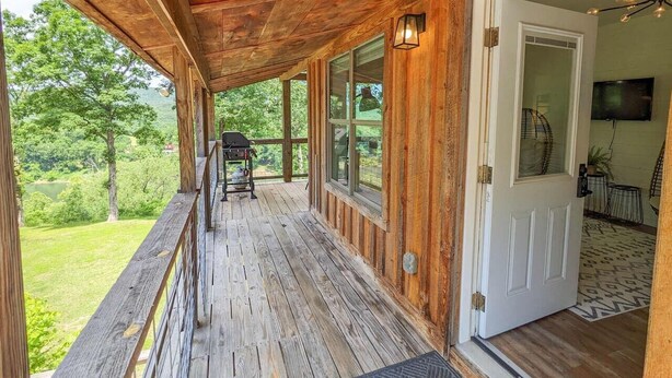 A wooden porch with a grill, a door leading inside, and a view of greenery. - The Crash Pad ~ Riverfront Retreat with Gameroom (Izard County)