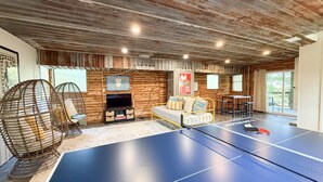 A ping pong table in a rustic room with wooden walls and ceiling, a sofa, and two wicker chairs. - The Crash Pad ~ Riverfront Retreat with Gameroom (Izard County)