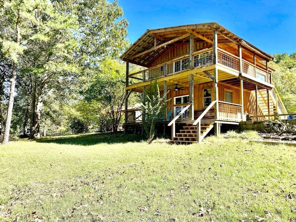 Una cabaña de madera de dos pisos con balcón, rodeada de árboles y un área con césped.