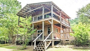 A multi-level wooden cabin with a balcony, surrounded by trees. - The Crash Pad ~ Riverfront Retreat with Gameroom (Izard County)