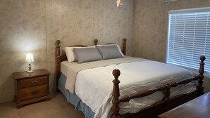 Une chambre à coucher avec un lit en bois, une table de chevet, une lampe, un ventilateur de plafond et une fenêtre avec des stores.