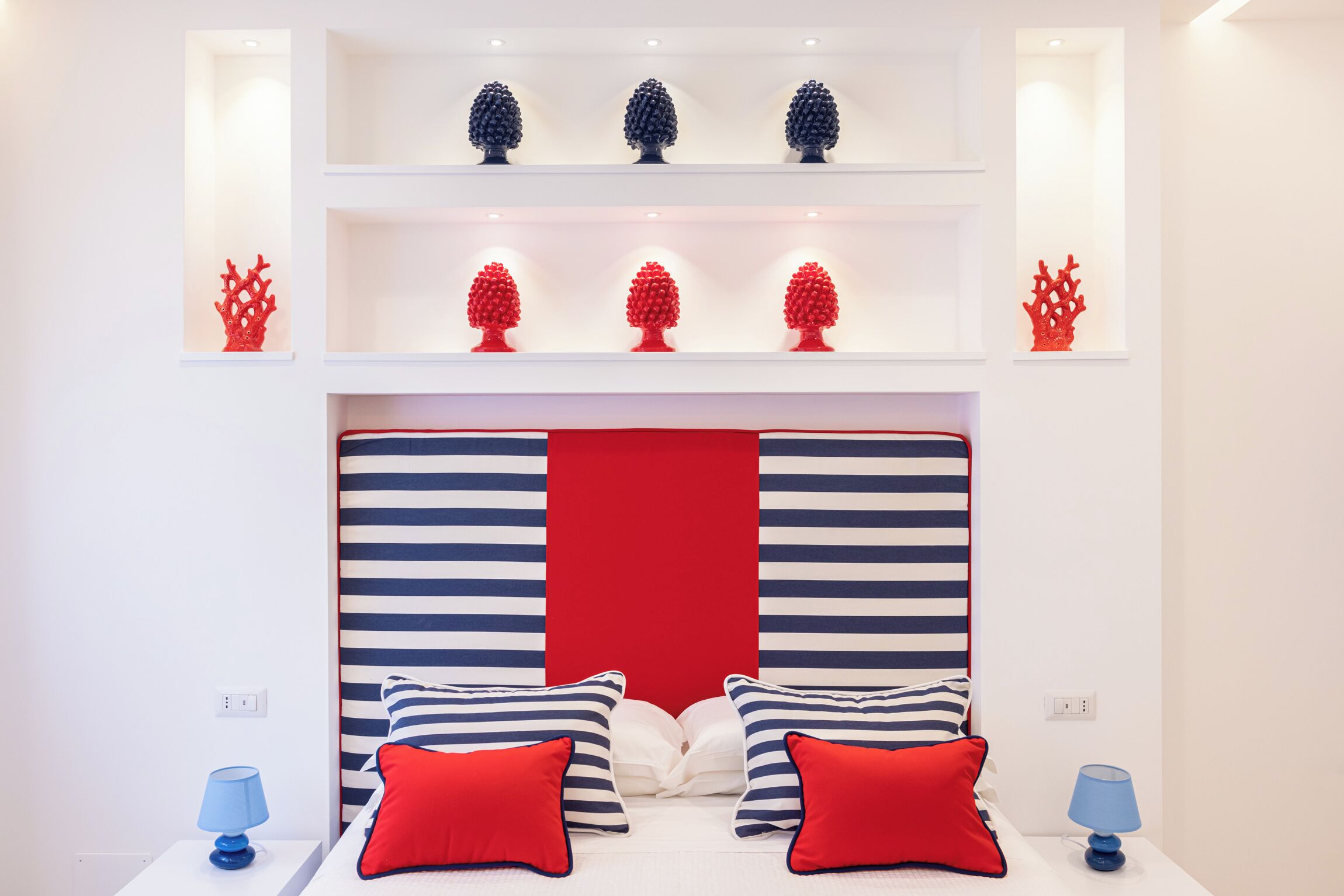 A hotel room with a striped headboard, red and white pillows, and decorative red and blue objects on shelves.