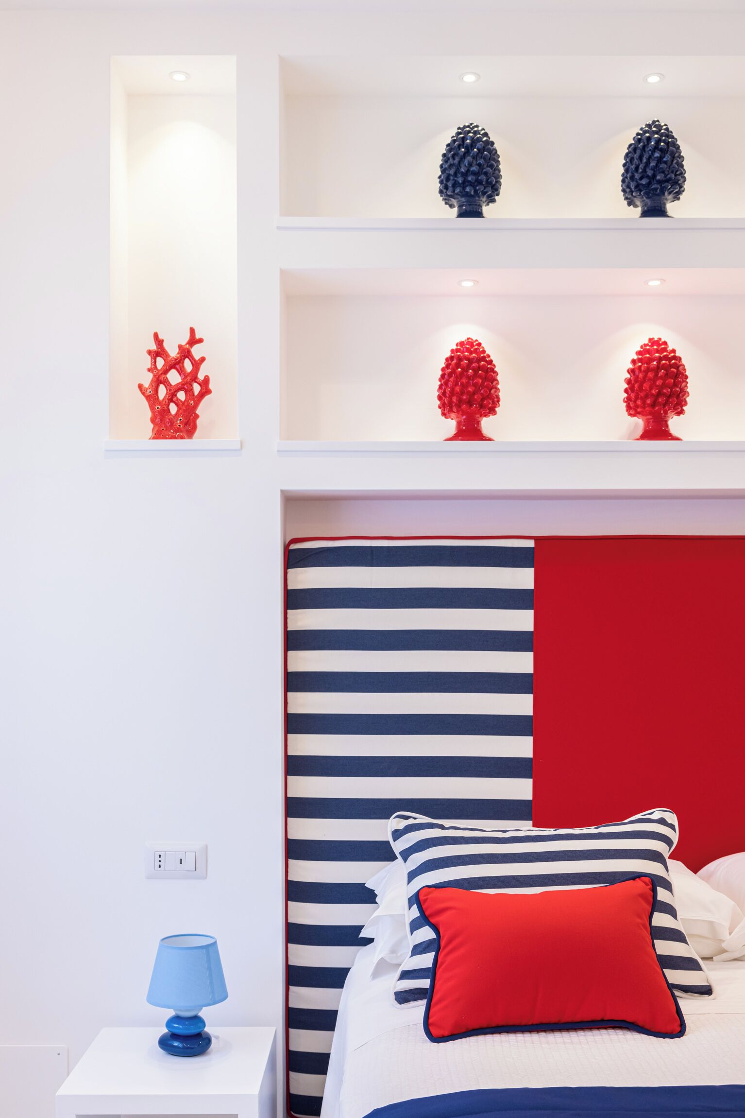 Un dormitorio moderno con una cabecera a rayas, almohadillas decorativas rojas y azules, y estantes empotrados con objetos esféricos rojos y azules.