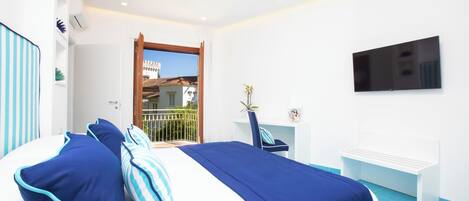 A modern hotel room with a bed, a desk, a chair, a TV, and a balcony with a view of buildings.