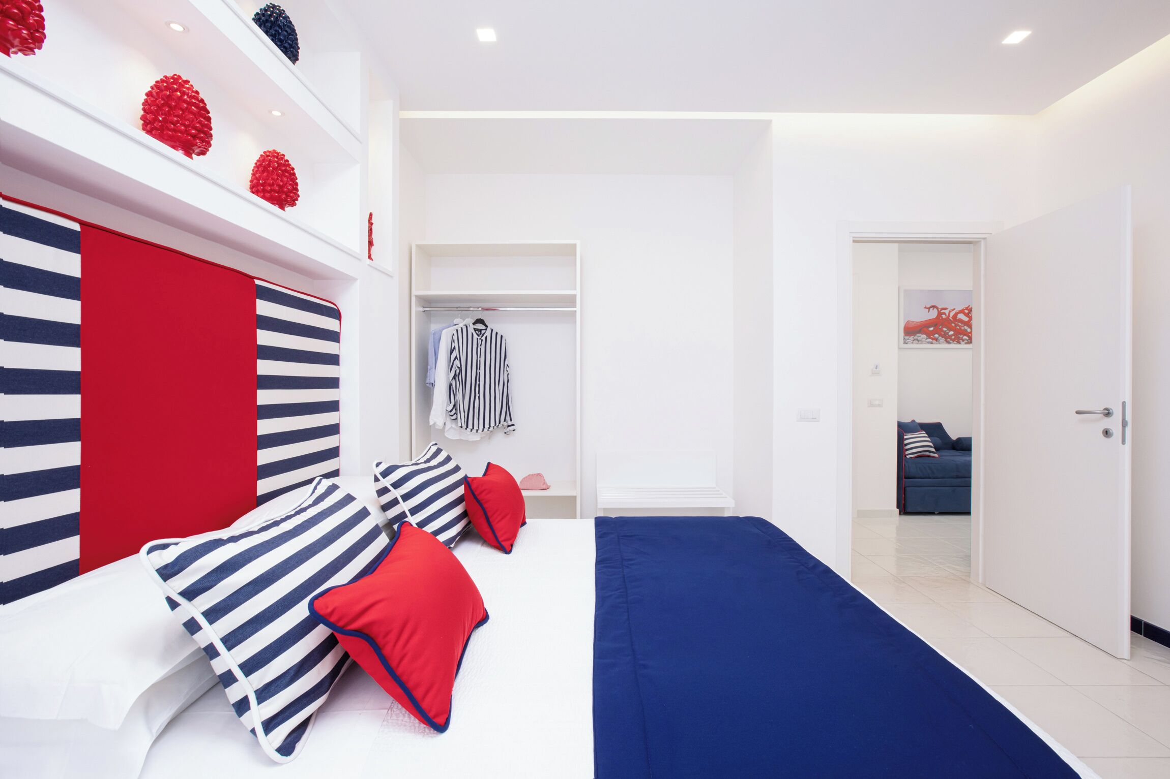 Une chambre à coucher moderne avec une tête de lit à rayures, un lit avec une literie rouge et bleue, un dressing et une porte donnant sur une autre pièce.