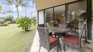 A patio with wicker furniture and a table, a glass door leading to a kitchen, and a view of palm trees. - Ground Floor condo suite with pool and ocean access (Kihei)