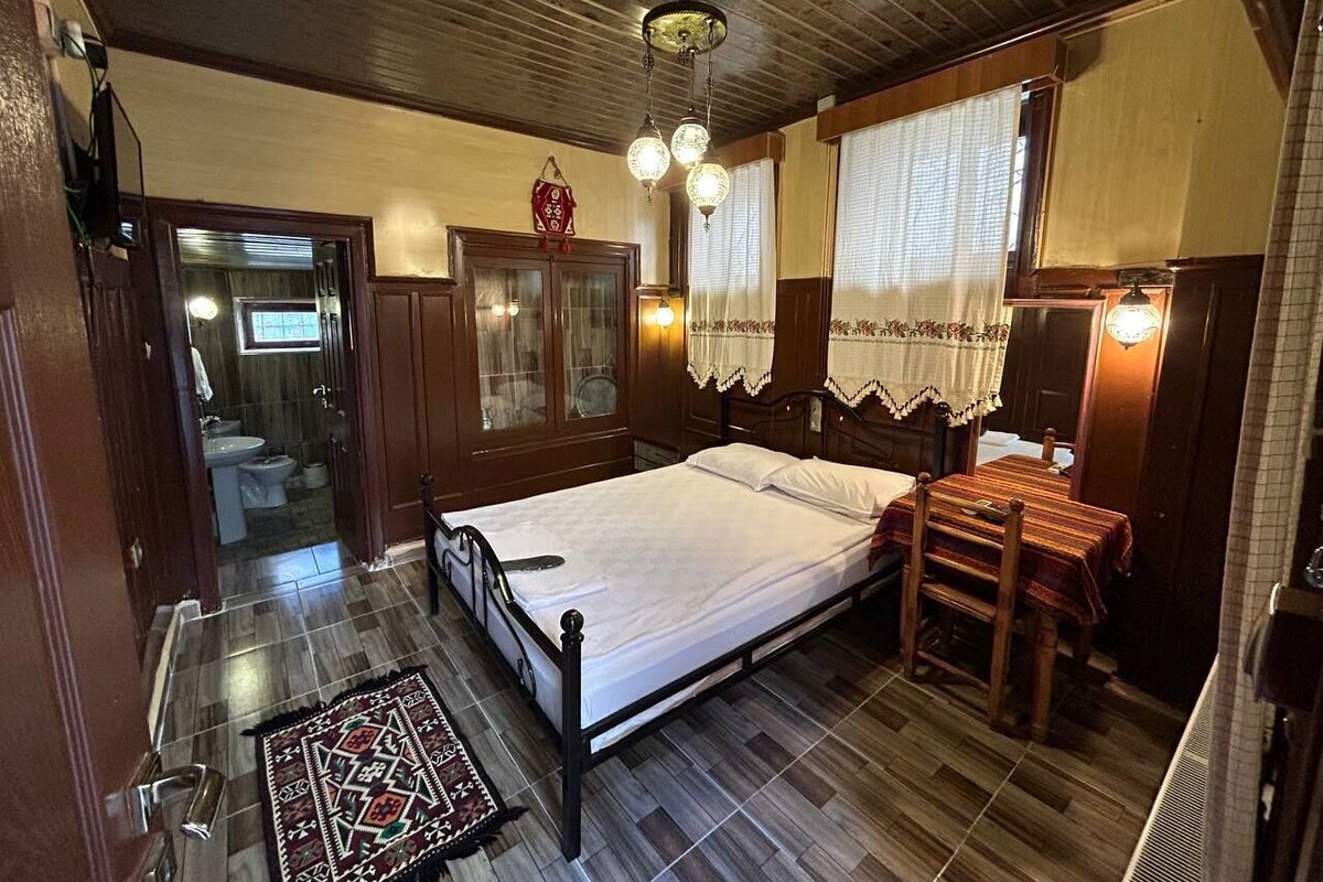 A bedroom with a wooden bed, a small table with chairs, a wooden wardrobe, and a window with curtains.