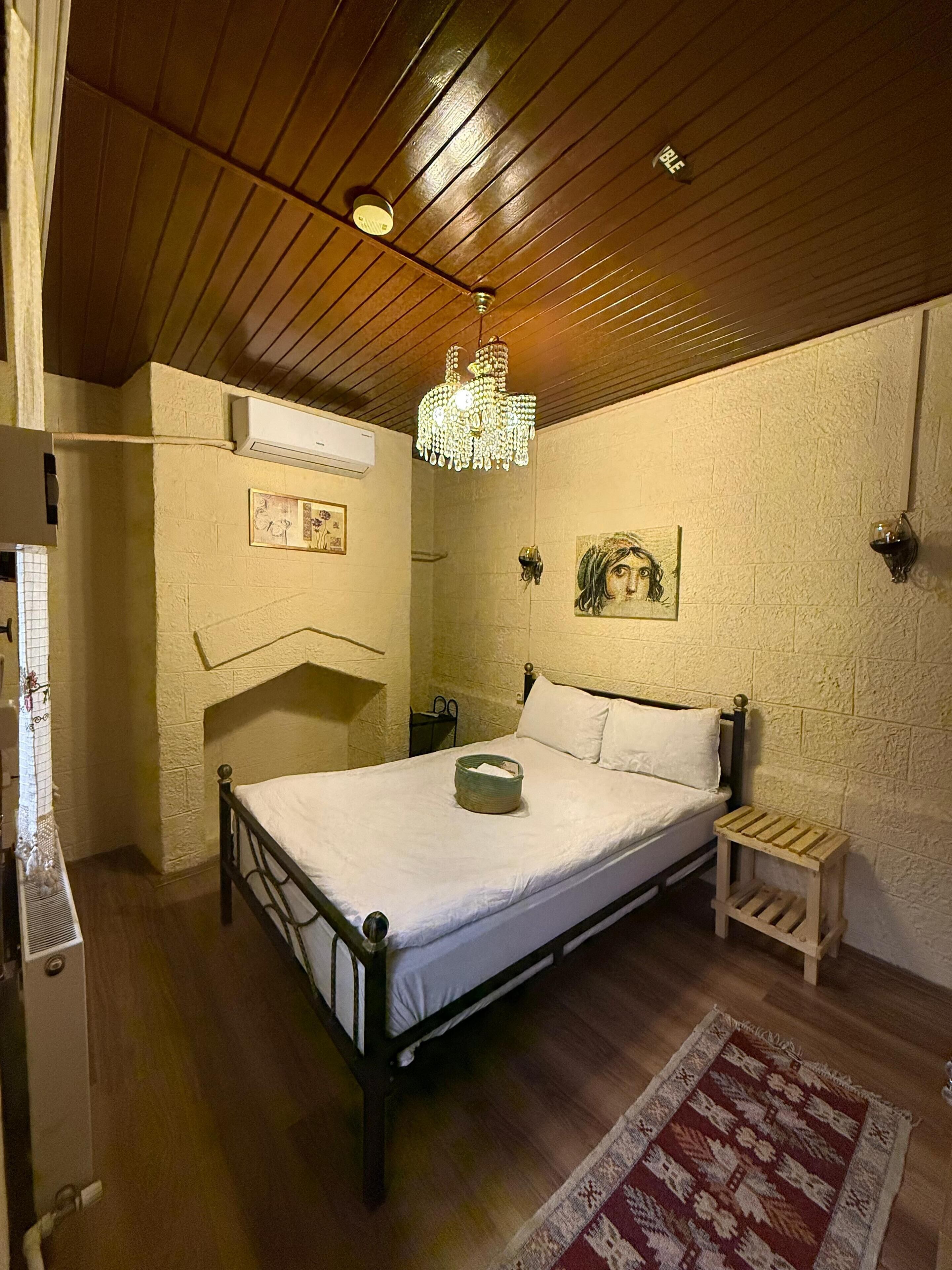 Un dormitorio con techo de madera, una cama con ropa de cama blanca, una mesita de noche pequeña, un aire acondicionado instalado en la pared y una alfombra estampada.