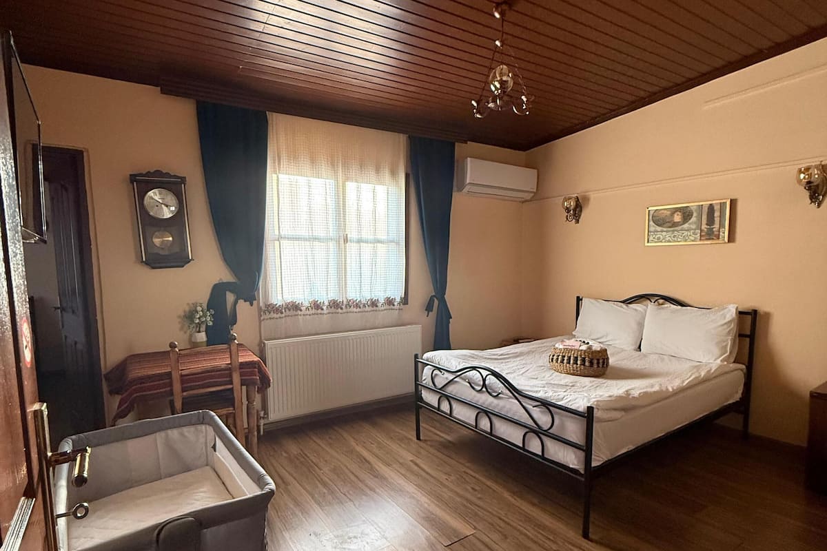 A bedroom with a wooden floor, a bed, a nightstand, a dresser, a window with curtains, an air conditioning unit, and a wall clock.