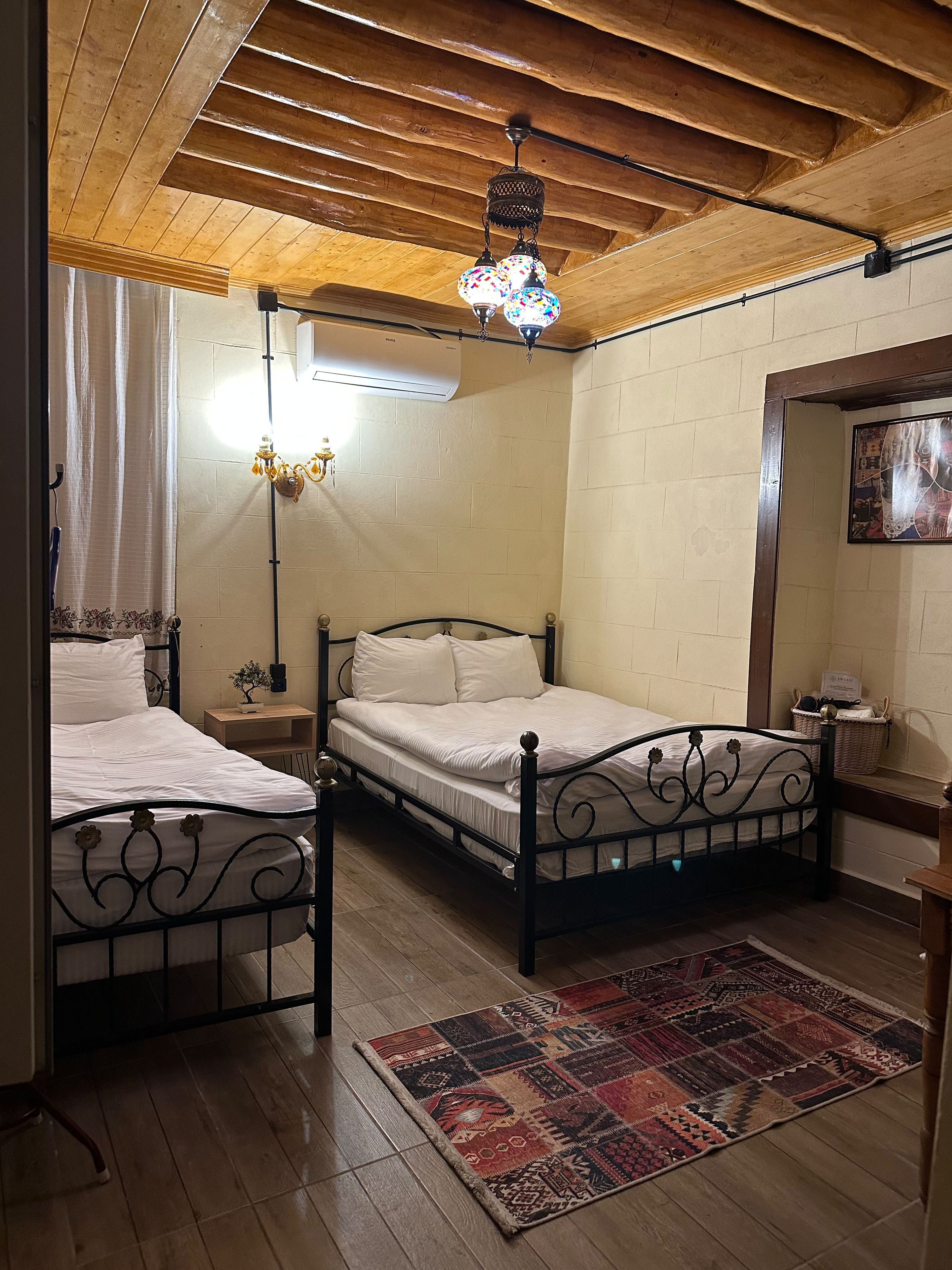 A bedroom with a double bed, a nightstand, a wall-mounted air conditioner, a patterned rug, and a wooden ceiling.