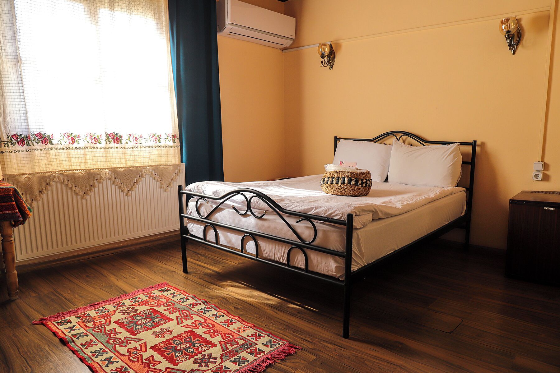 A single bed with a metal frame, a bedside table, a small basket, and a red patterned rug on a wooden floor.
