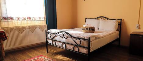 A single bed with a metal frame, a bedside table, a small basket, and a red patterned rug on a wooden floor.