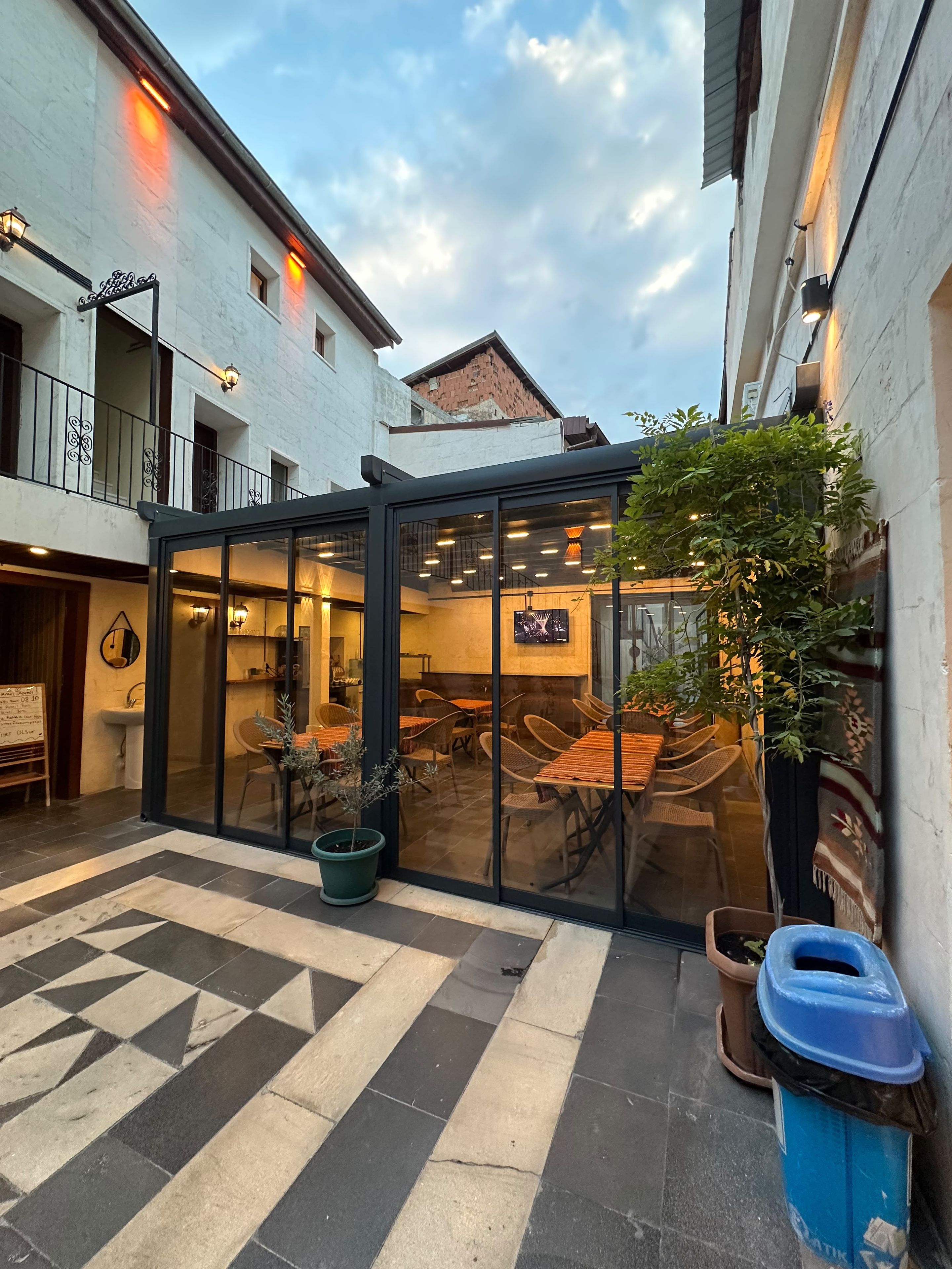 A courtyard with a glass-enclosed dining area, potted plants, and a trash bin.