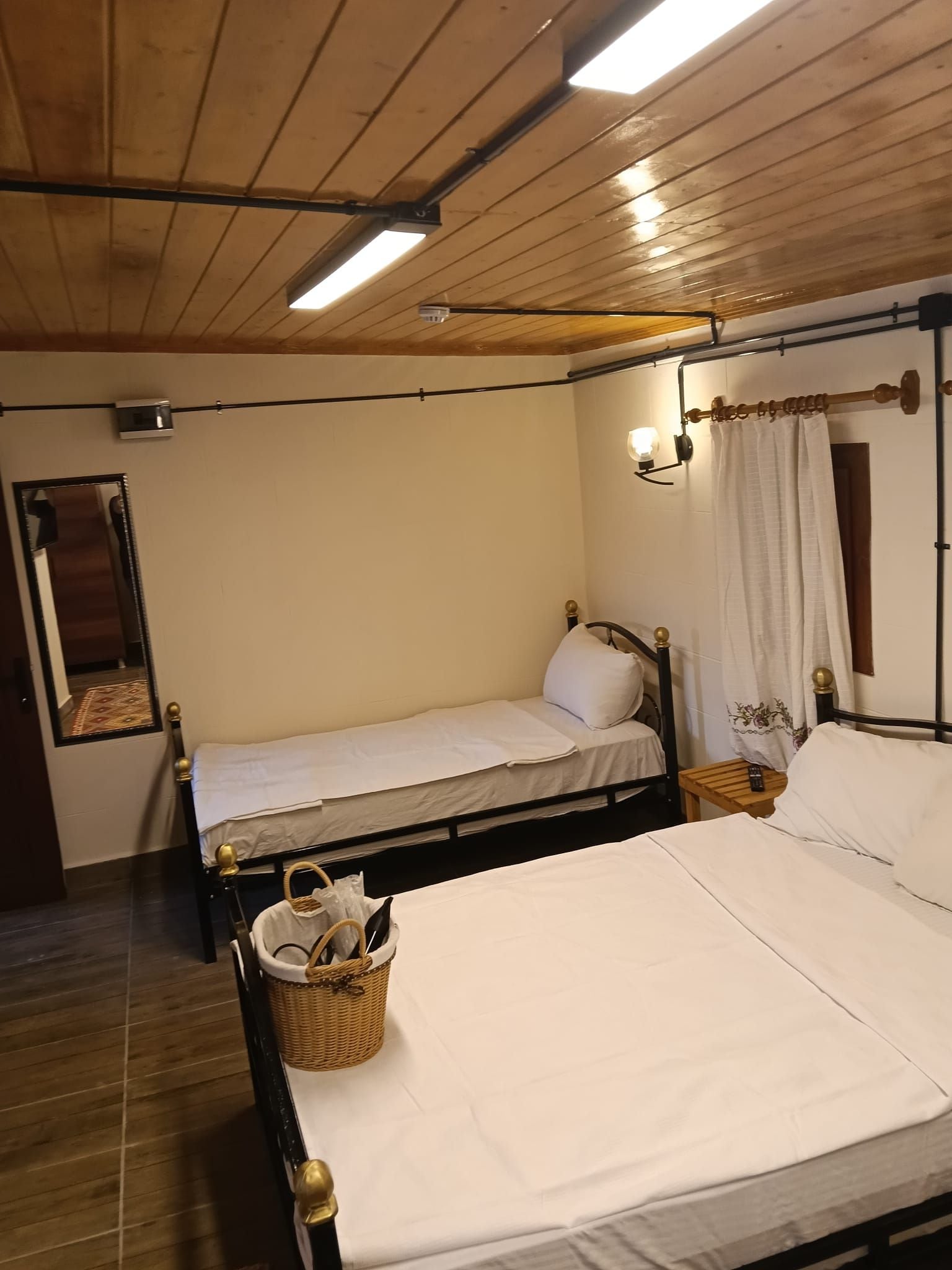 Un dormitorio con dos camas individuales, techo de madera, un espejo, una lámpara de pared y una ventana con cortinas.