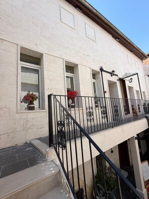 Ein Gebäude mit weißer Fassade, zahlreichen Fenstern, einem Balkon mit schwarzen Geländern und einem kleinen Topfpflanzen auf der Fensterbank.