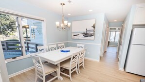 A dining area with a white table and chairs, a chandelier, and a beach scene painting on the wall. - Oceanside, Lighthouse Views, Community Pool, Community Amenities, 601, Our Happy Place (Corolla)