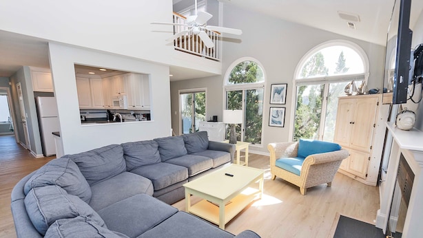 A spacious living room with a grey sectional sofa, a small coffee table, and a wicker chair. There is a kitchen in the background with white cabinets and a ceiling fan. - Oceanside, Lighthouse Views, Community Pool, Community Amenities, 601, Our Happy Place (Corolla)