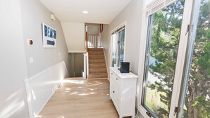 A hallway with a wooden floor, white walls, a white radiator, a white cabinet with a TV, and a large window with a view of trees. - Oceanside, Lighthouse Views, Community Pool, Community Amenities, 601, Our Happy Place (Corolla)