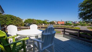 A deck with a round table and chairs, overlooking a park with colorful houses and a clear sky. - Oceanside, Lighthouse Views, Community Pool, Community Amenities, 601, Our Happy Place (Corolla)