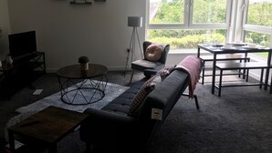 Una sala de estar con sofá, mesa de centro, televisor y una zona de comedor con mesa y sillas. Hay cuadros en la pared y una ventana con vistas a zonas verdes.