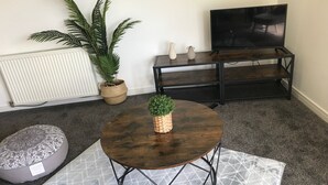 Una sala de estar con una mesa de centro redonda de madera, un otomán gris con estampado, una planta en maceta, un televisor sobre un soporte negro y un radiador.